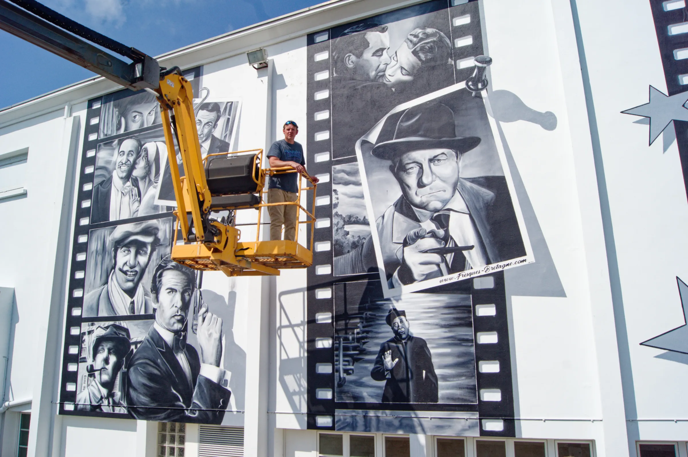 Fresque monumentale de portraits d'acteurs en noir et blanc sur pellicules de cinéma, peinte depuis une nacelle sur la façade de la salle de cinéma de Plouescat